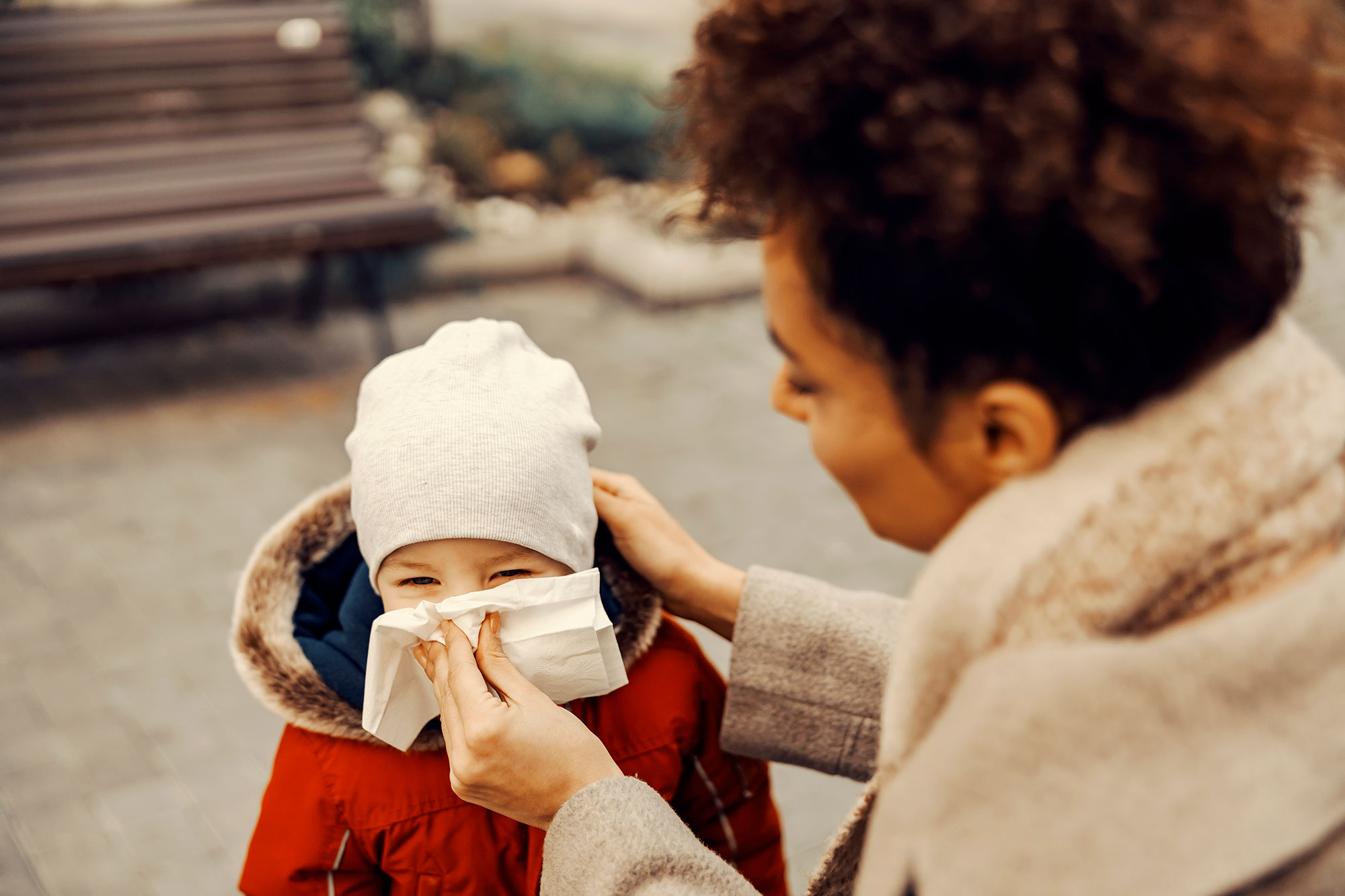 A woman blowing the nose of a little boy in a park during cold weather.