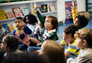 A group of happy kids in kindergarten class sitting on the floor. Some are raising their hands to answer or ask a question.
