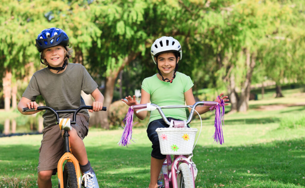 A young brother and sister riding bikes together in a park-like setting and wearing bike helmets for safety.