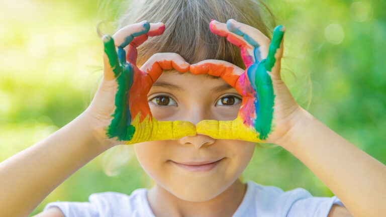A young girl with multi color paint on her hands makes a heart shape over her face.