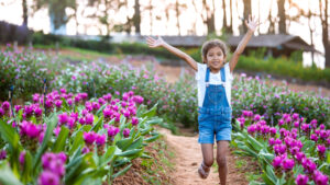 Smiling young girl in denim overalls and a white tee shirt runs joyfully through a spring flower field surrounded by blooming purple tulips—capturing the spirit of outdoor family fun in Portland, Oregon.