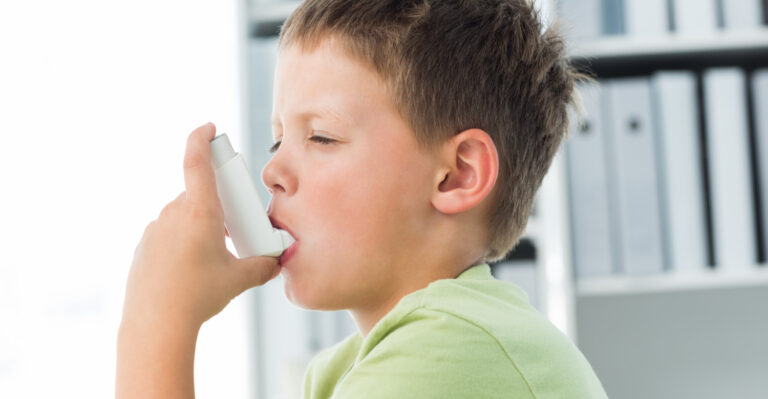 A young boy in a green shirt uses an asthma inhaler, inhaling medication to manage his symptoms. This image accompanies a blog about recognizing and managing asthma in children.