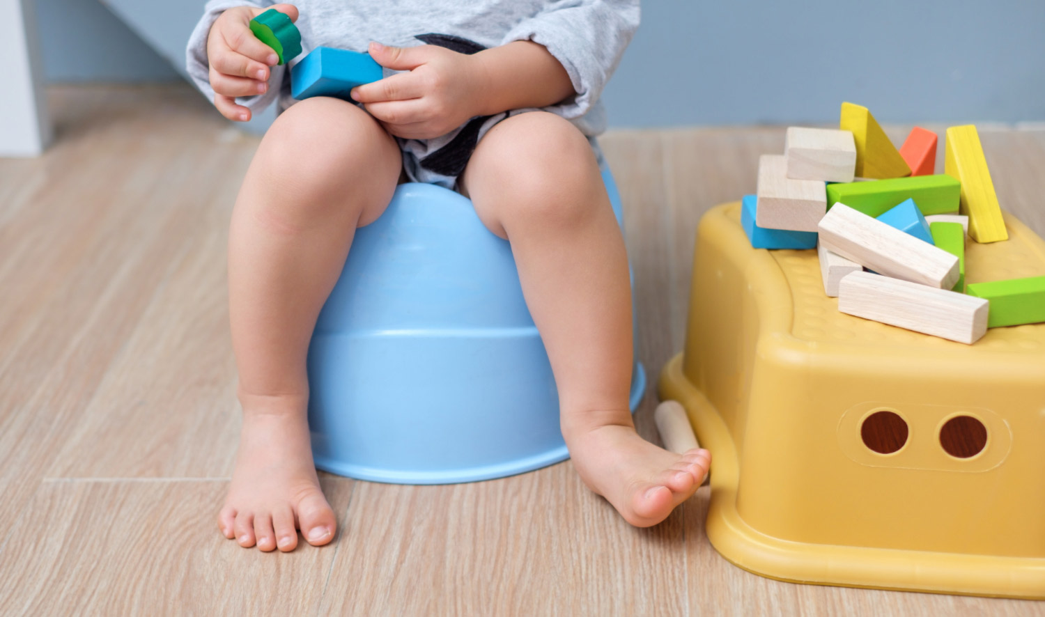 A toddler sits on a blue potty seat holding colorful toy blocks, with more blocks stacked on a yellow step stool nearby. This image accompanies a pediatrician-written blog offering tips and tricks for potty training toddlers.