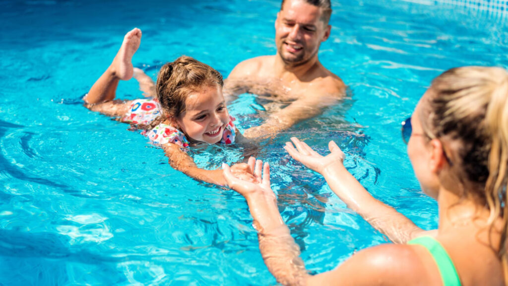 A young girl and parents are in a bright blue pool learning how to swim.