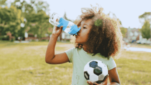 A young black girl with a soccer ball drinks from water bottle on a hot day, emphasizing the importance of recognizing and preventing dehydration in kids.