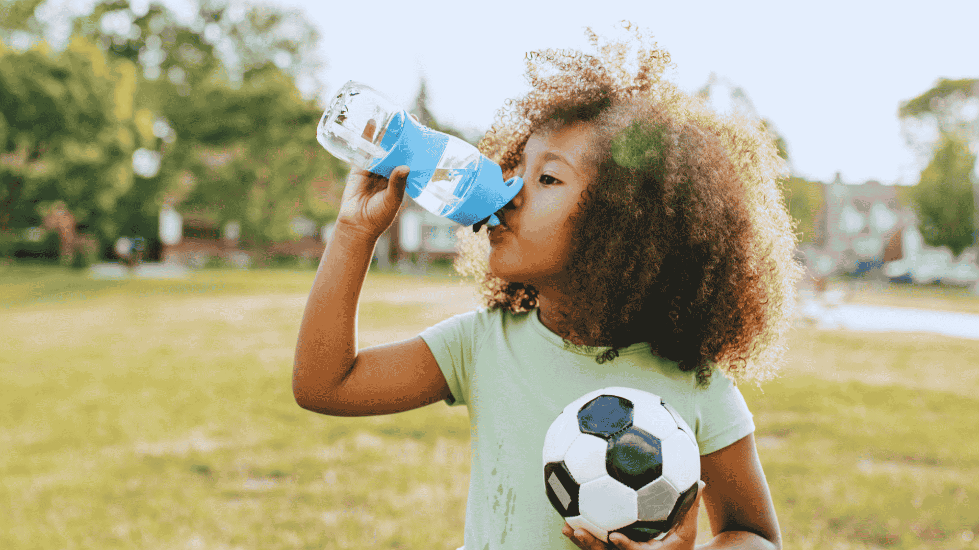 A young black girl with a soccer ball drinks from water bottle on a hot day, empasizing the importance of recognizing and preventing dehydration in kids.