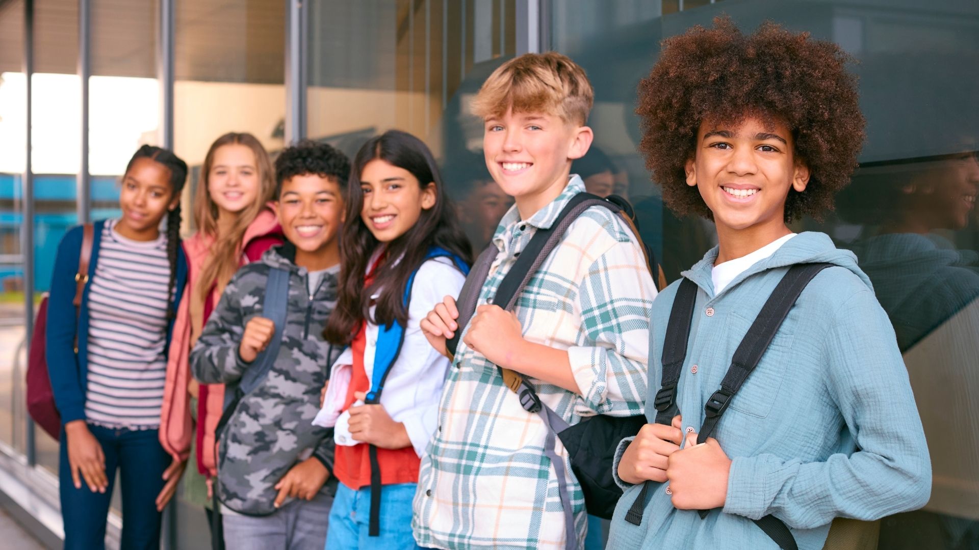 A group of diverse high school students wearing backpacks stand outside a school building in front of lockers.