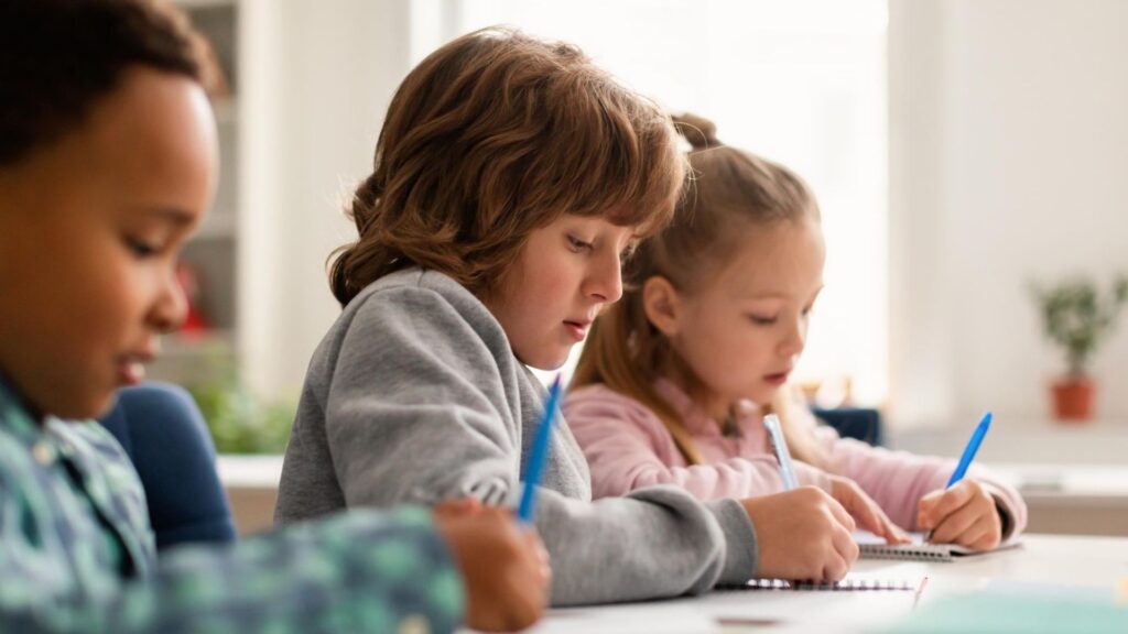 Three young children sit at a classroom table focused on writing in their notebooks. The setting is bright and calm, suggesting a school environment. This image represents focus and learning—common challenges and goals discussed in blogs about ADHD symptoms and diagnosis.