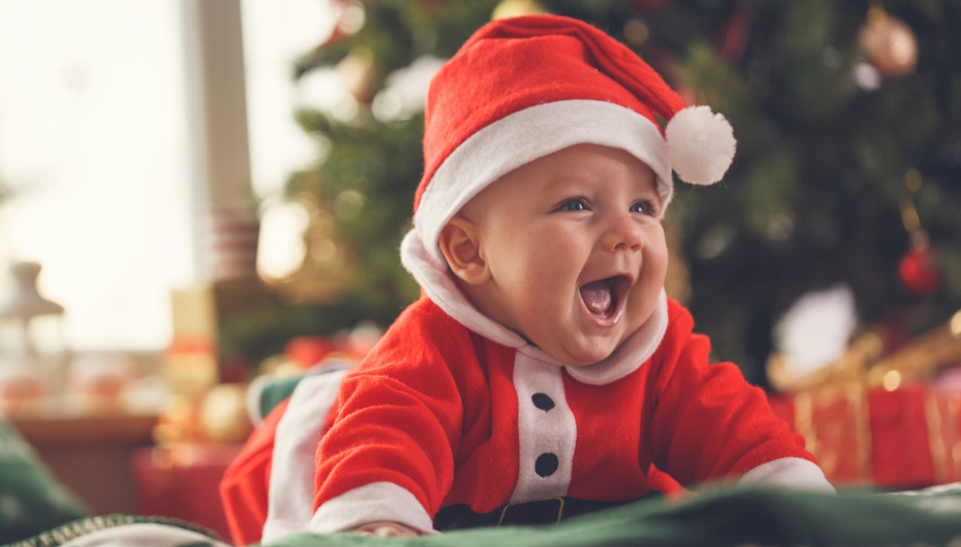 Smiling baby dressed in a Santa outfit lying on their stomach in front of a decorated Christmas tree, part of a blog about preventing holiday injuries with tips for parents and caregivers.