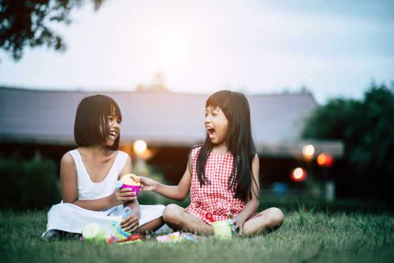 Two young girls sitting on the grass outdoors laughing and playing together during a playdate, sharing toys and enjoying time with a friend.