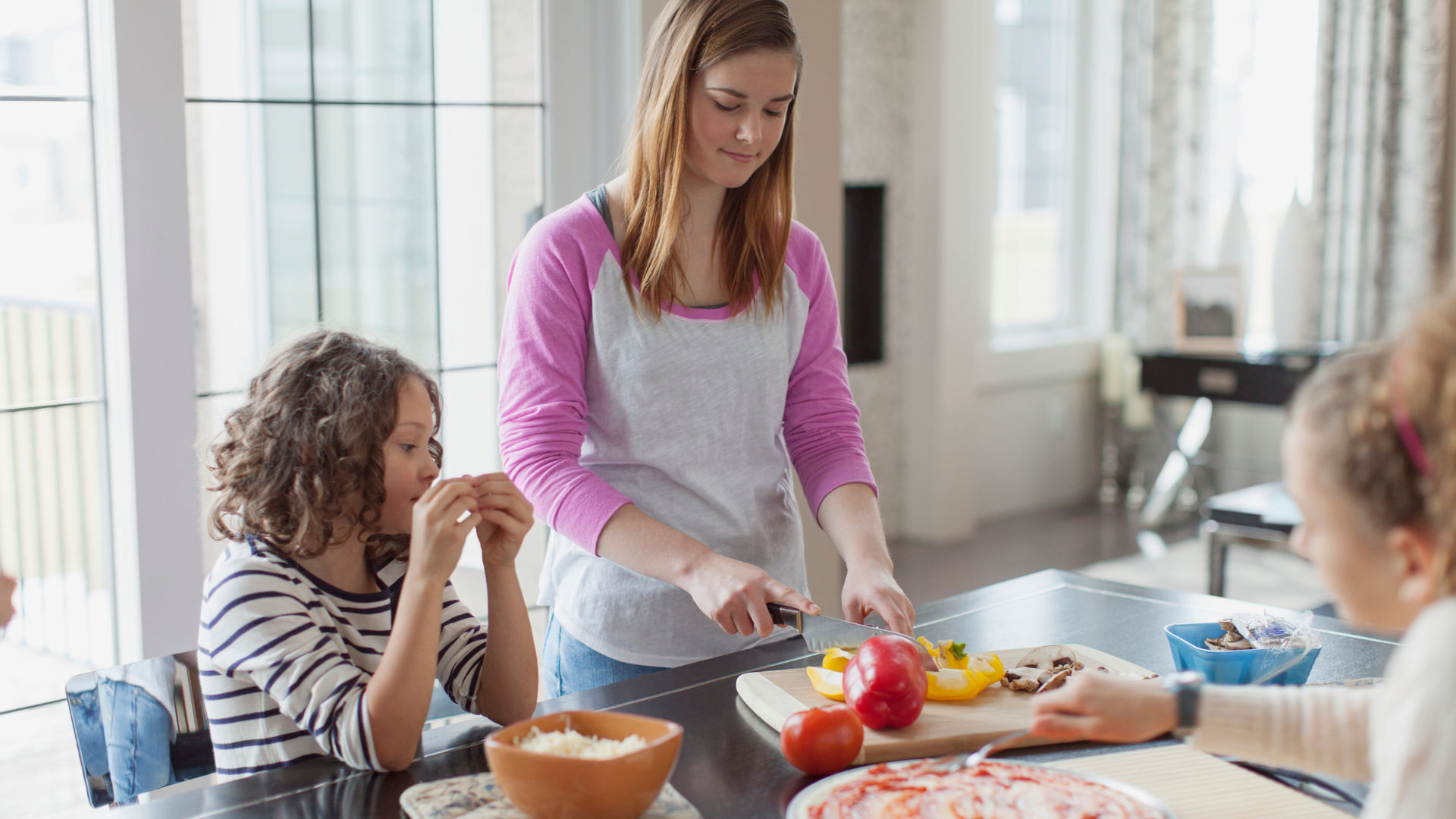 Babysitter prepares a meal in a bright kitchen, slicing vegetables on a cutting board while two children sit nearby—one watching closely and another seated at the table as food is served.